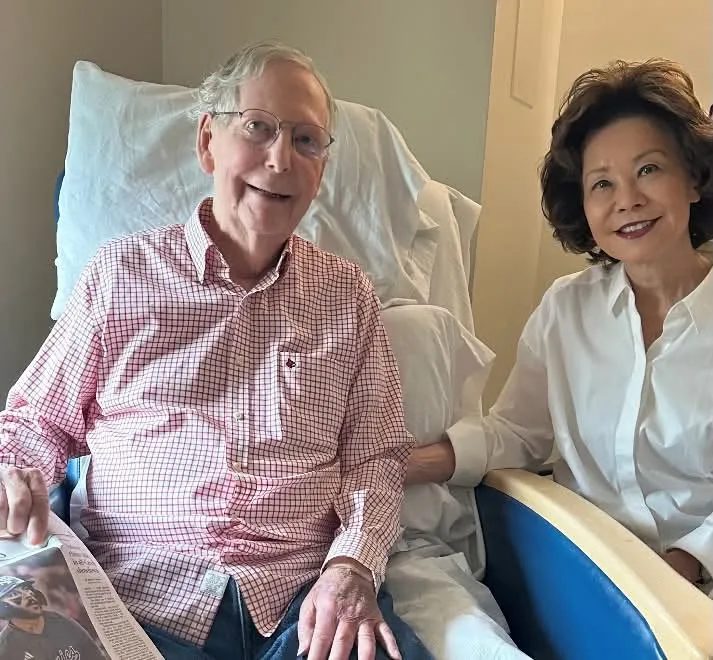 Photo released by Sen. McConnell's office showing the senator in a hospital bed holding a newspaper, with Elaine Chao beside him. Credit: Office of Sen. Mitch McConnell